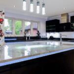 Large striking feature island in black gloss with marble effect quartz worktop and handle trim LED lighting framed by bubble tube pendant lighting above.